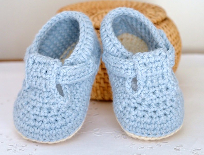 May include: A pair of light blue crocheted baby shoes with a white trim. The shoes have a t-strap design and are made with a soft, textured yarn.