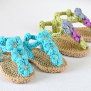 May include: Three pairs of crocheted baby sandals with flower embellishments. The sandals are in shades of blue, green, and purple.
