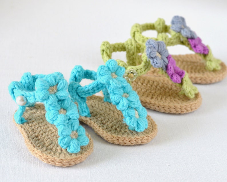May include: Three pairs of crocheted baby sandals with flower embellishments. The sandals are in shades of blue, green, and purple.