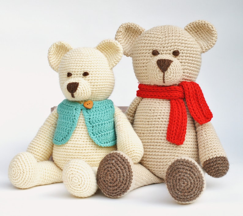 May include: Two handmade crochet teddy bears. One bear wears a teal vest with a wooden button, the other a red scarf. Both have brown noses and eyes, and tan bodies. The bears are sitting, with brown-colored feet.