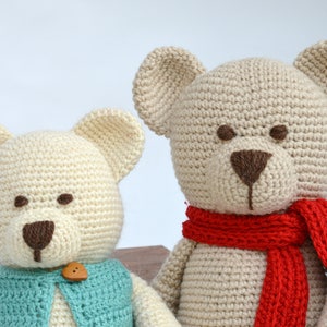 May include: Two handmade crochet teddy bears. One bear wears a blue vest with a wooden button, the other has a red scarf. Both have cream-colored bodies, brown eyes, and noses.