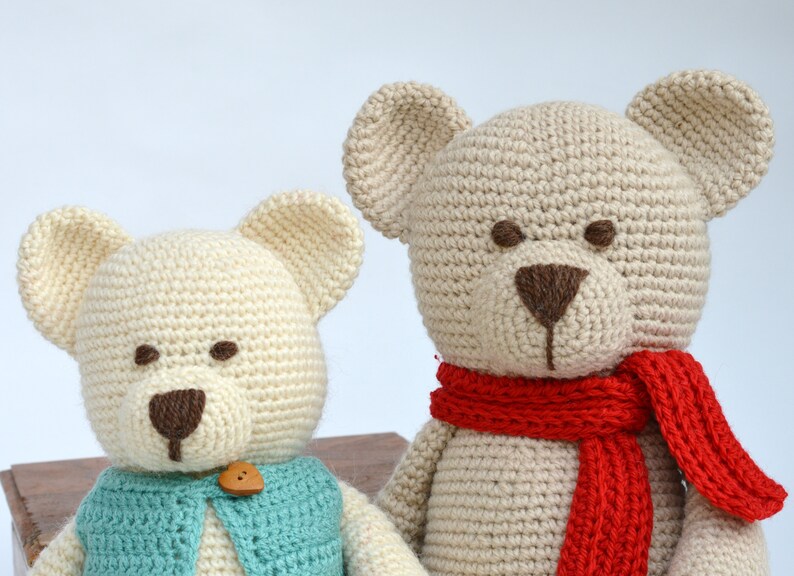 May include: Two handmade crochet teddy bears. One bear wears a blue vest with a wooden button, the other has a red scarf. Both have cream-colored bodies, brown eyes, and noses.