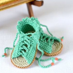 K&ouml;nnte beinhalten: Ein Paar geh&auml;kelte Babysandalen in hellgr&uuml;n mit braunem Besatz. Die Sandalen haben Schn&uuml;rsenkel, die vorne gebunden werden.