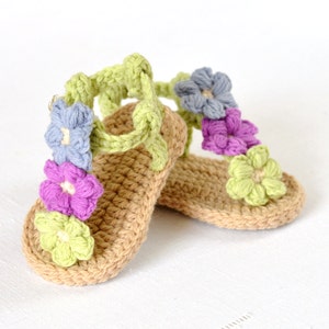 May include: A pair of crocheted baby sandals with a light brown base and colourful flower embellishments. The flowers are in shades of green, purple, blue, and light pink.