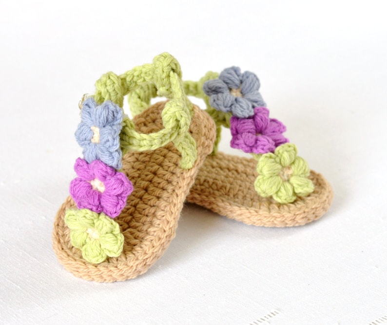 May include: A pair of crocheted baby sandals with a light brown base and colourful flower embellishments. The flowers are in shades of green, purple, blue, and light pink.