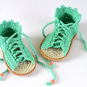 K&ouml;nnte beinhalten: Ein Paar gr&uuml;ne geh&auml;kelte Babysandalen mit braunen Sohlen und Schn&uuml;rsenkeln. Die Sandalen haben einen gewellten Rand und sind mit einer Schleife gebunden.