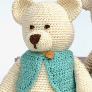 May include: A handmade crochet teddy bear with a cream-colored body, brown eyes and nose, and a turquoise vest. The vest is fastened with a wooden heart-shaped button. The bear has large, rounded ears.