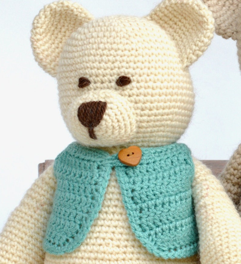 May include: A handmade crochet teddy bear with a cream-colored body, brown eyes and nose, and a turquoise vest. The vest is fastened with a wooden heart-shaped button. The bear has large, rounded ears.