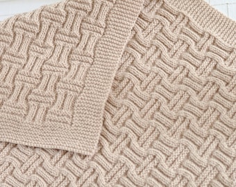Op de afbeelding: Een close-up van een beige gebreide deken met een geweven patroon. De deken is dubbelgevouwen, waardoor de ingewikkelde details van het breiwerk zichtbaar zijn.
