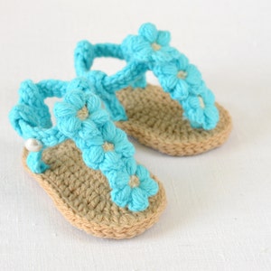 May include: A pair of crocheted baby sandals with blue flower embellishments. The sandals have a tan sole and a white pearl accent.