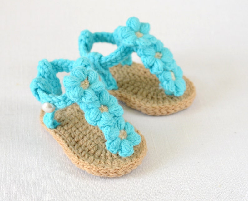 May include: A pair of crocheted baby sandals with blue flower embellishments. The sandals have a tan sole and a white pearl accent.
