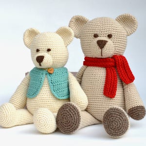 May include: Two handmade crochet teddy bears. One bear wears a teal vest with a wooden button, the other a red scarf. Both have brown noses and paws, and are cream-colored.
