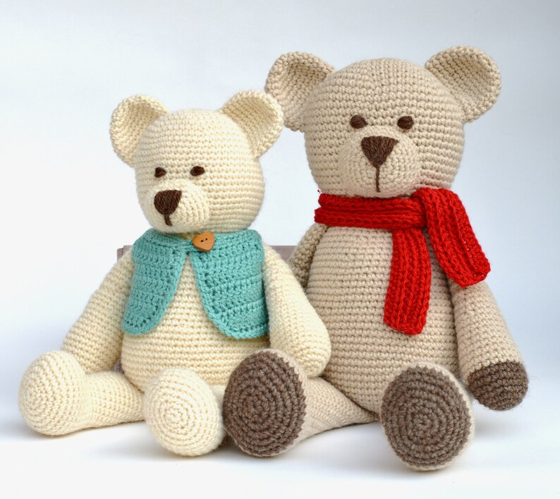 May include: Two handmade crochet teddy bears. One bear wears a teal vest with a wooden button, the other a red scarf. Both have brown noses and paws, and are cream-colored.