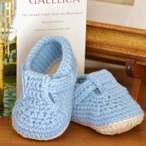 May include: A pair of light blue crocheted baby shoes with white trim. The shoes have a simple design with a strap across the top.