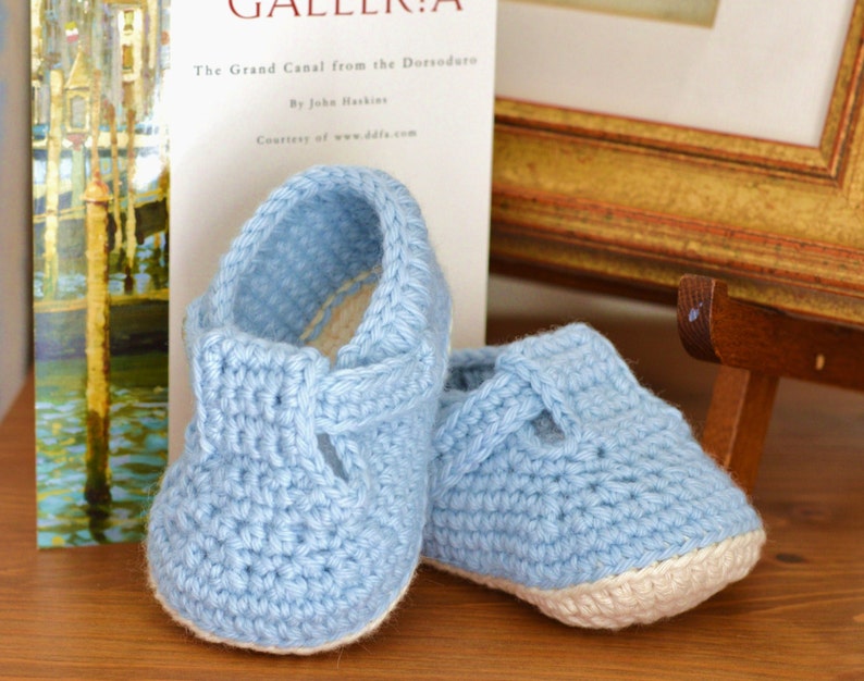 May include: A pair of light blue crocheted baby shoes with white trim. The shoes have a simple design with a strap across the top.