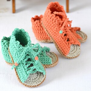 K&ouml;nnte beinhalten: Zwei Paar geh&auml;kelte Babysandalen. Ein Paar ist gr&uuml;n und das andere orange. Beide Paare haben eine braune geh&auml;kelte Sohle.