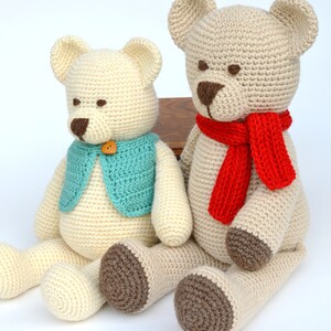 May include: Two handmade crochet teddy bears. One bear wears a teal vest with a wooden button. The other bear has a red scarf. Both bears are cream-colored with brown accents on the nose and feet. They are sitting on a white surface.