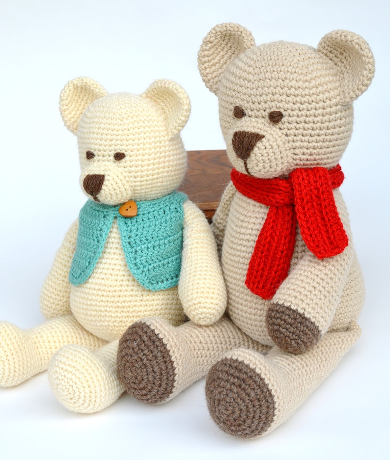 May include: Two handmade crochet teddy bears. One bear wears a teal vest with a wooden button. The other bear has a red scarf. Both bears are cream-colored with brown accents on the nose and feet. They are sitting on a white surface.