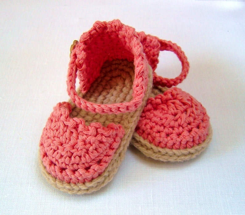 May include: A pair of crocheted baby shoes in pink and beige. The shoes have a strap that goes around the ankle and a buckle closure.