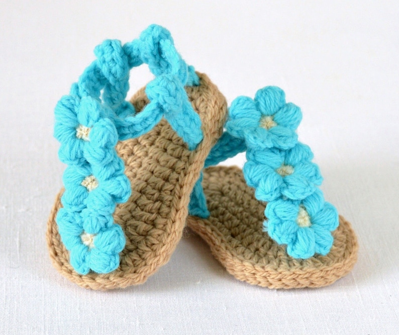 May include: A pair of crocheted baby sandals with blue flower embellishments. The sandals are made of brown yarn and have a strap that goes around the ankle.