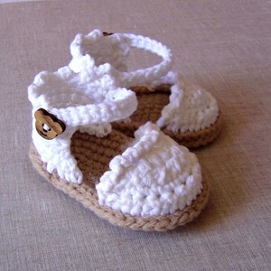May include: White crocheted baby sandals with brown button accents. The sandals have a brown woven sole.