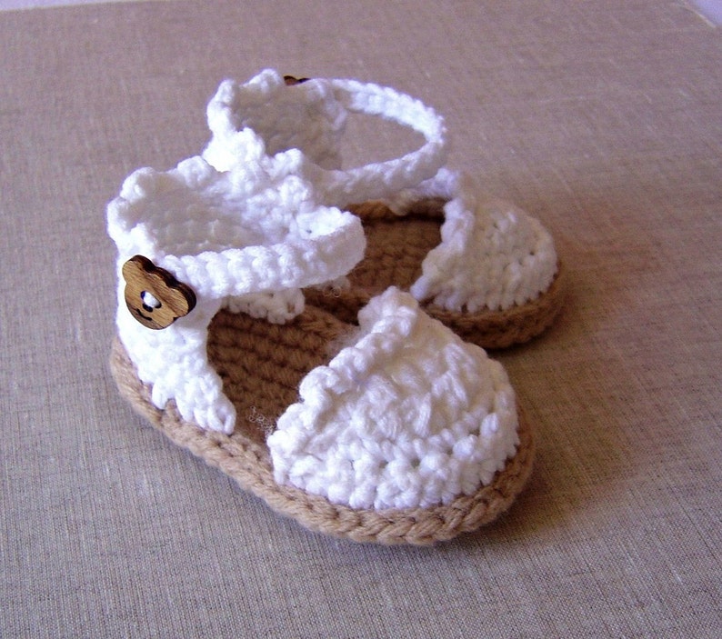 May include: White crocheted baby sandals with brown button accents. The sandals have a brown woven sole.