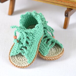 K&ouml;nnte beinhalten: Ein Paar geh&auml;kelte Babysandalen in hellgr&uuml;n mit braunen Verzierungen. Die Sandalen haben ein Schn&uuml;rdesign und sind aus weichem, bequemem Garn gefertigt.