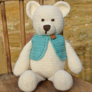 May include: A handmade, cream-colored crochet teddy bear with a turquoise vest and a brown button. The bear has brown eyes and a brown nose. It is sitting on a wooden chair. The bear is a soft toy.