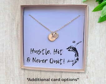 May include: A gold necklace with a volleyball charm and a small round charm. The necklace is in a white box with a black and white illustration of a volleyball player and the text "Hustle. Hit & Never Quit!"