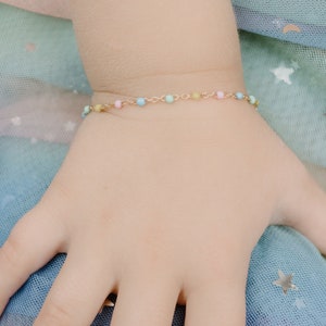 May include: A delicate gold bracelet with small, pastel-coloured beads in pink, blue, and yellow. The bracelet is on a child's wrist, against a soft, multi-coloured background.