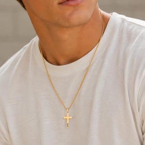 May include: A gold-toned metal cross pendant suspended on a thin, matching gold-toned chain. The cross features a vertical orientation with a smooth, polished finish and a small, dark engraved detail visible on the lower portion of the cross bar.