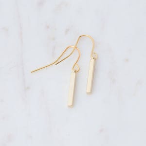 May include: A pair of gold-toned drop earrings featuring slender, rectangular bar pendants suspended from curved ear wires. The earrings have a smooth, polished finish and a simple, linear design that hangs vertically from the delicate metal hooks.