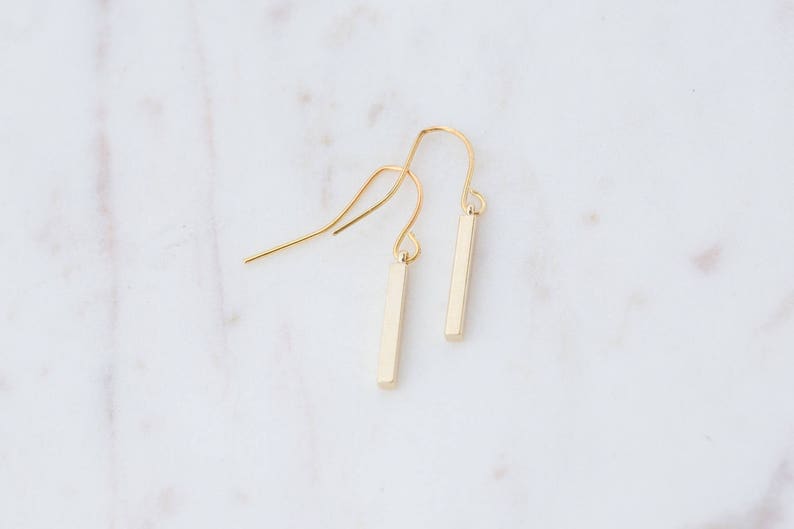 May include: A pair of gold-toned drop earrings featuring slender, rectangular bar pendants suspended from curved ear wires. The earrings have a smooth, polished finish and a simple, linear design that hangs vertically from the delicate metal hooks.
