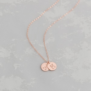 Puede incluir: Un collar de oro rosa con dos colgantes redondos, cada uno grabado con una letra. Las letras son "A" y "M".