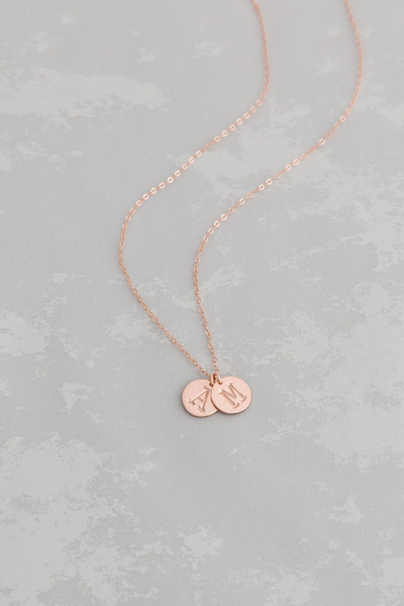 Puede incluir: Un collar de oro rosa con dos colgantes redondos, cada uno grabado con una letra. Las letras son "A" y "M".