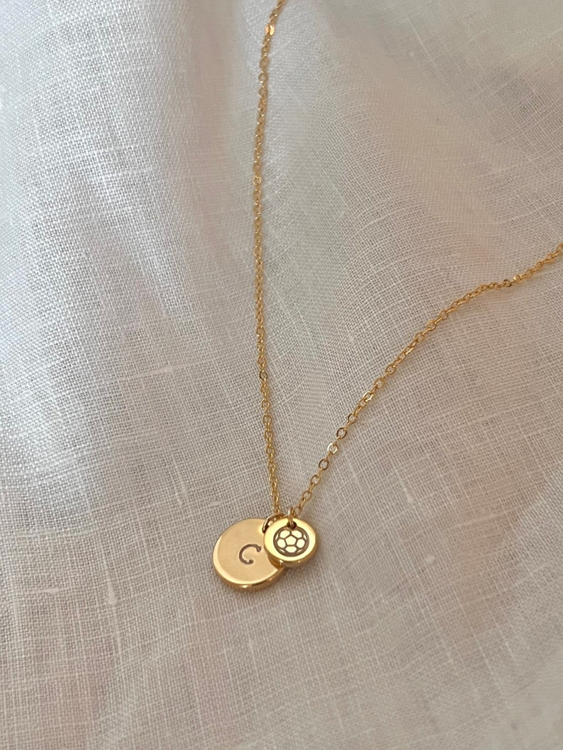 May include: A gold chain necklace with two charms. One charm is a round disc with the letter "C" engraved on it. The other charm is a round disc with a soccer ball design.