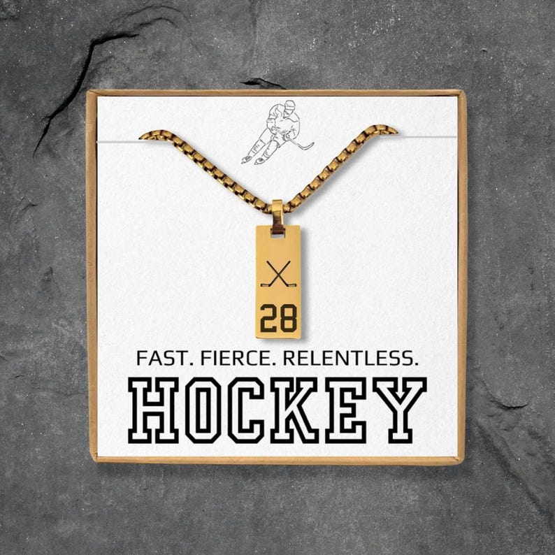 May include: A gold-colored hockey pendant necklace with a rectangular tag featuring crossed hockey sticks and the number 28. The necklace is presented in a white box with the word "HOCKEY" and the text "FAST. FIERCE. RELENTLESS."