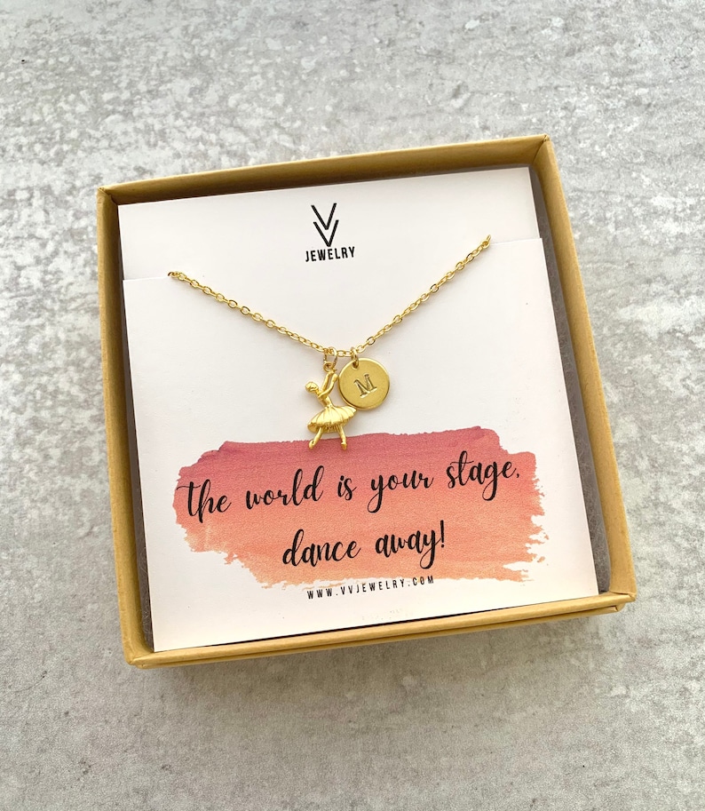 May include: Gold necklace with a ballerina charm and a personalized initial disc. The necklace comes in a box with a watercolor background and the text "The world is your stage. Dance away!" and the website address "www.vvjewelry.com".