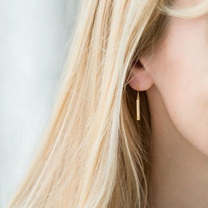 May include: A gold-toned metal earring featuring a simple, slender cylindrical bar pendant suspended from a delicate hook wire. The earring hangs vertically, showcasing a smooth, polished finish that reflects light along its straight, minimalist form.