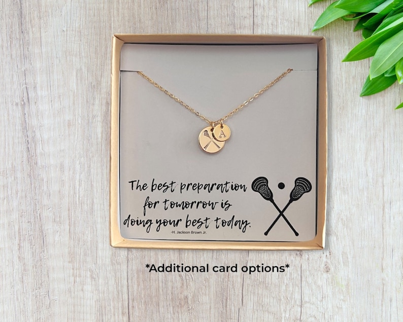 May include: A gold necklace with a lacrosse stick charm and a letter 'A' charm in a gift box. The box has a quote that reads "The best preparation for tomorrow is doing your best today." - H. Jackson Brown Jr.