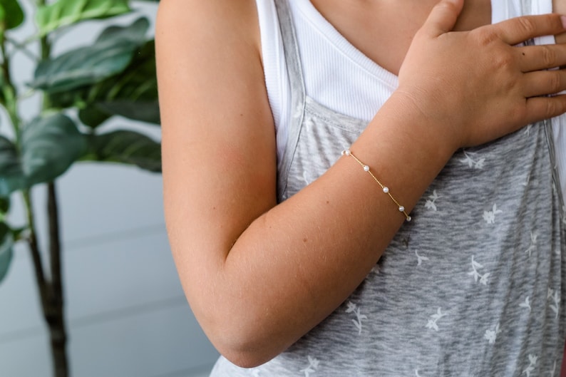 May include: A delicate gold bracelet with small, white pearls is worn on a person's wrist. The bracelet is strung with several pearls and is set against the person's skin and a grey vest top.