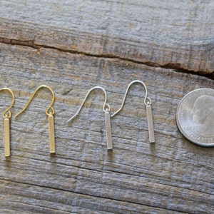 May include: Two pairs of drop earrings featuring thin, vertical rectangular bars suspended from curved ear wires. One pair is gold-toned and the other is silver-toned. A United States quarter coin is placed next to the earrings for scale.