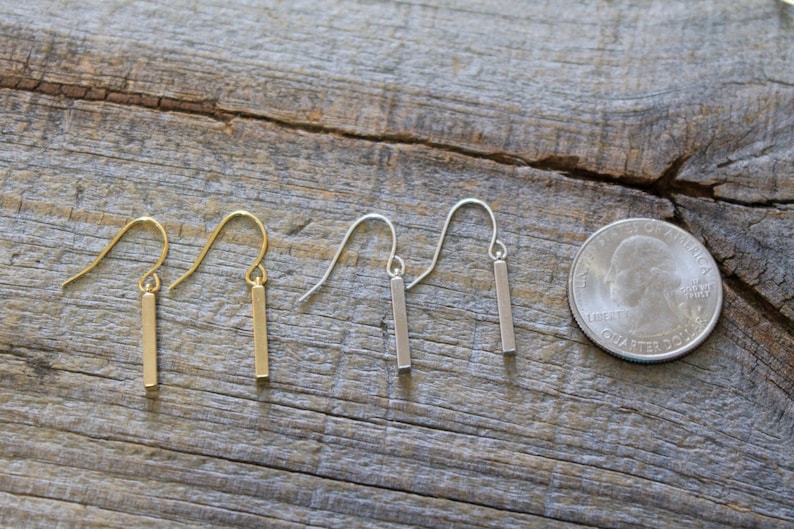 May include: Two pairs of drop earrings featuring thin, vertical rectangular bars suspended from curved ear wires. One pair is gold-toned and the other is silver-toned. A United States quarter coin is placed next to the earrings for scale.