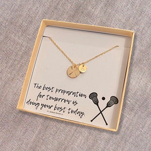 May include: A gold necklace with a lacrosse stick charm and a letter 'A' charm in a white box with the quote "The best preparation for tomorrow is doing your best today." - H. Jackson Brown Jr.