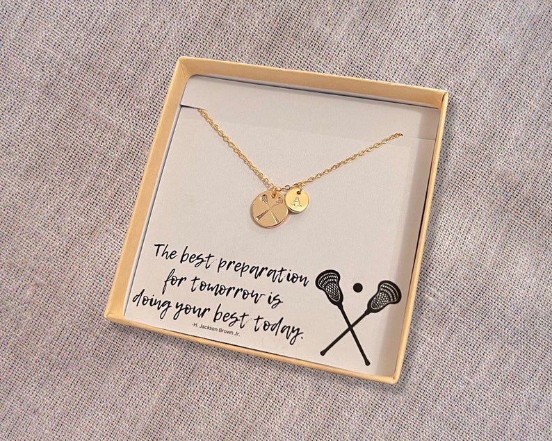 May include: A gold necklace with a lacrosse stick charm and a letter 'A' charm in a white box with the quote "The best preparation for tomorrow is doing your best today." - H. Jackson Brown Jr.