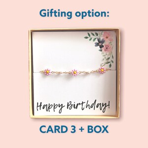 May include: A gold chain bracelet with pink daisy charms, presented in a gift box. The card inside reads "Happy Birthday!" with floral accents. The box is gold-coloured and the text "CARD 3 + BOX" is at the bottom.