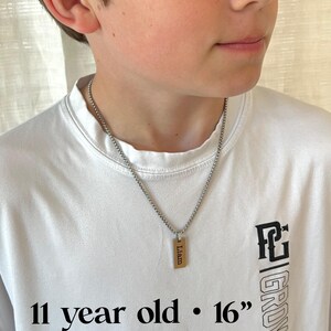 May include: A silver chain necklace with a rectangular pendant engraved with the name "Liam". The pendant is gold-colored. The necklace is approximately 16 inches long. The person is wearing a white t-shirt with black text that says "11 year old" and "GROW".