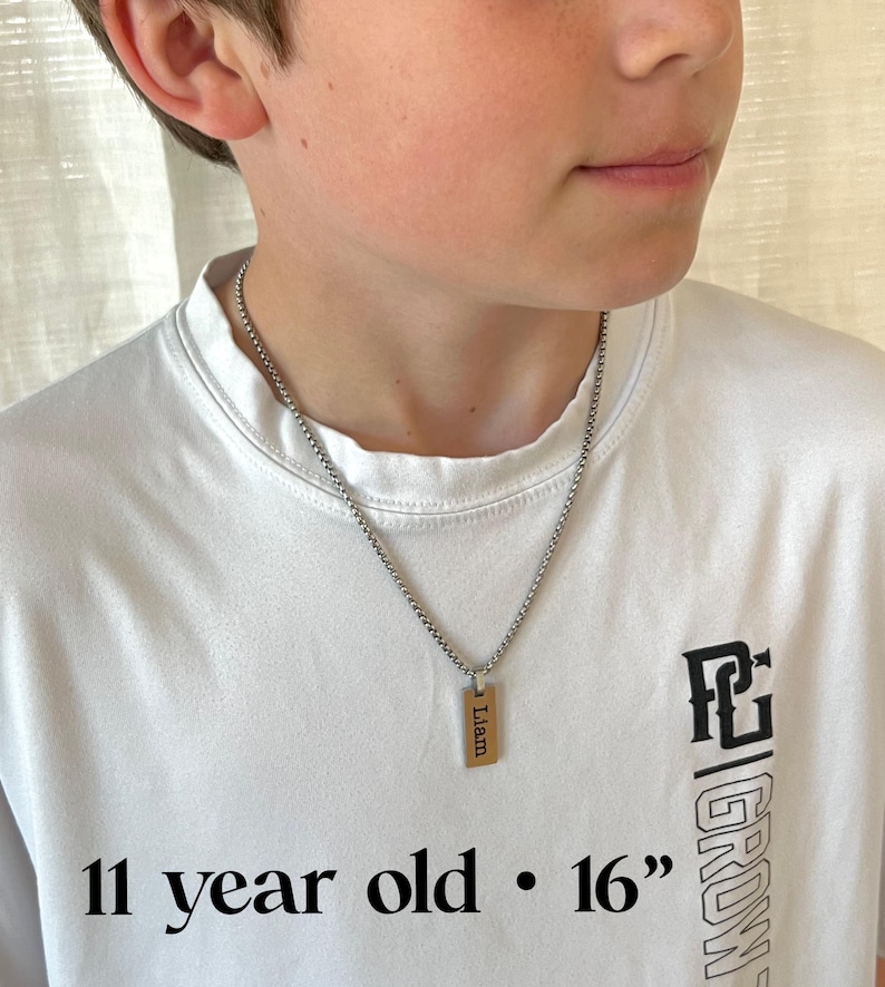 May include: A silver chain necklace with a rectangular pendant engraved with the name "Liam". The pendant is gold-colored. The necklace is approximately 16 inches long. The person is wearing a white t-shirt with black text that says "11 year old" and "GROW".