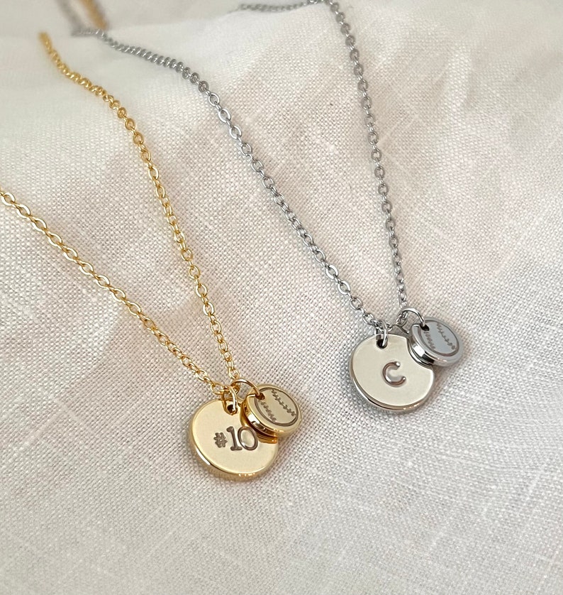May include: Two gold and silver necklaces with baseball charms. One necklace has a charm with the number 10 and the other has a charm with the letter C.