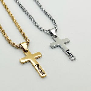 May include: Two cross necklaces. One is gold-colored with the name "Bennett" engraved on it. The other is silver-colored with the name "Liam" engraved. Both necklaces have a rope chain.